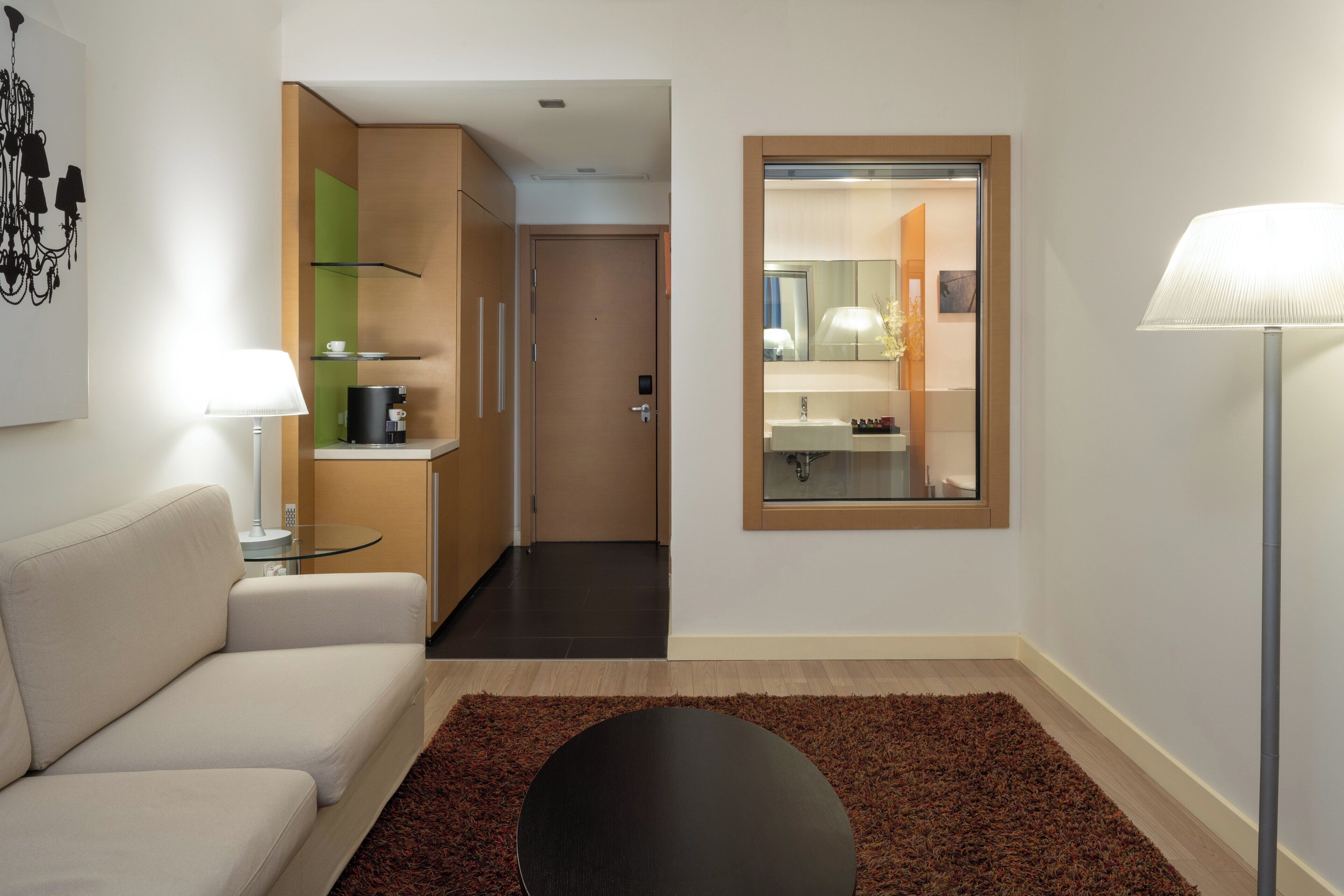 A modern hotel room with a sofa, a round table, a coffee machine, a mirror, and a lamp.