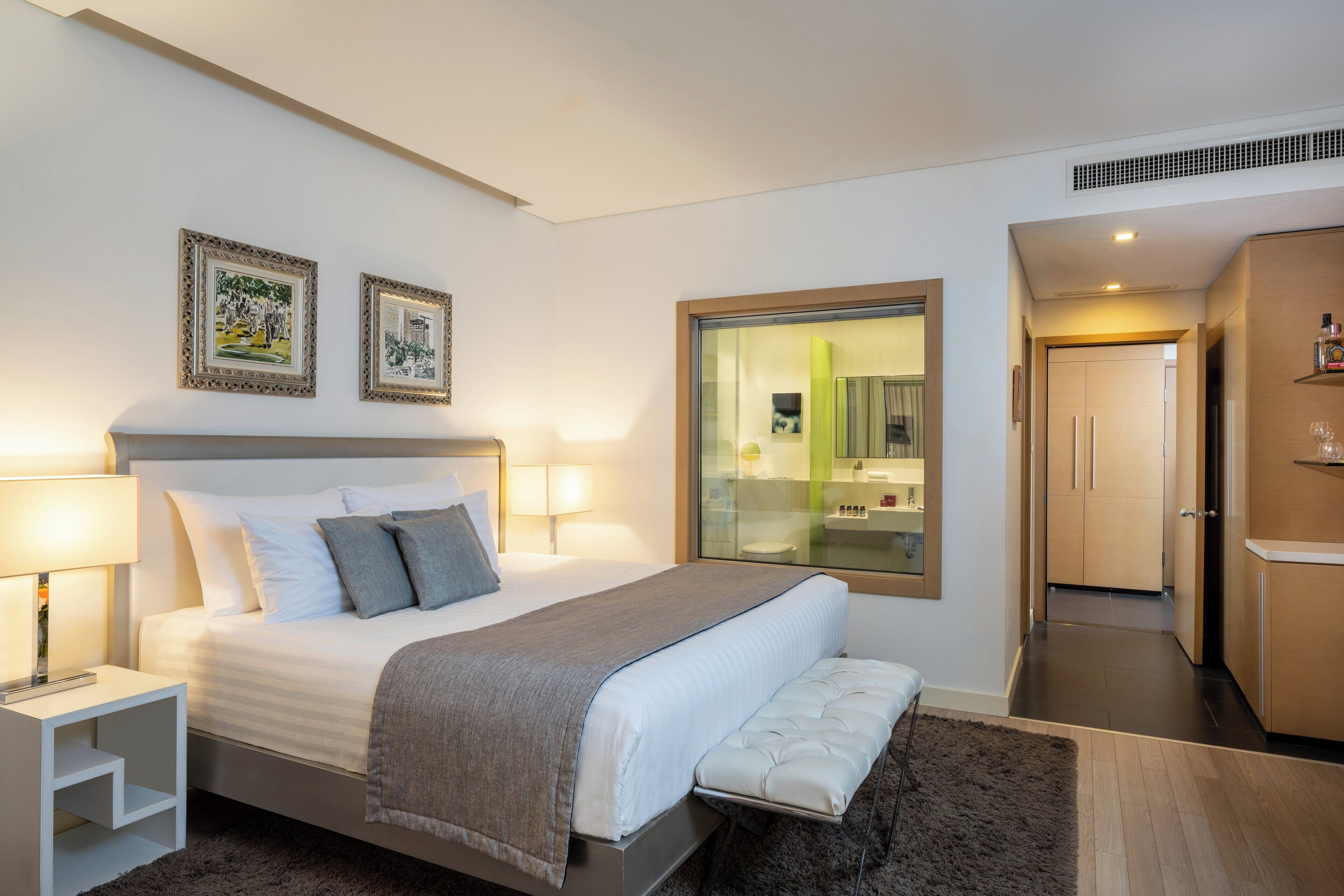 A modern hotel room with a large bed, bedside lamps, a bench, a glass wall, and a view of the bathroom.