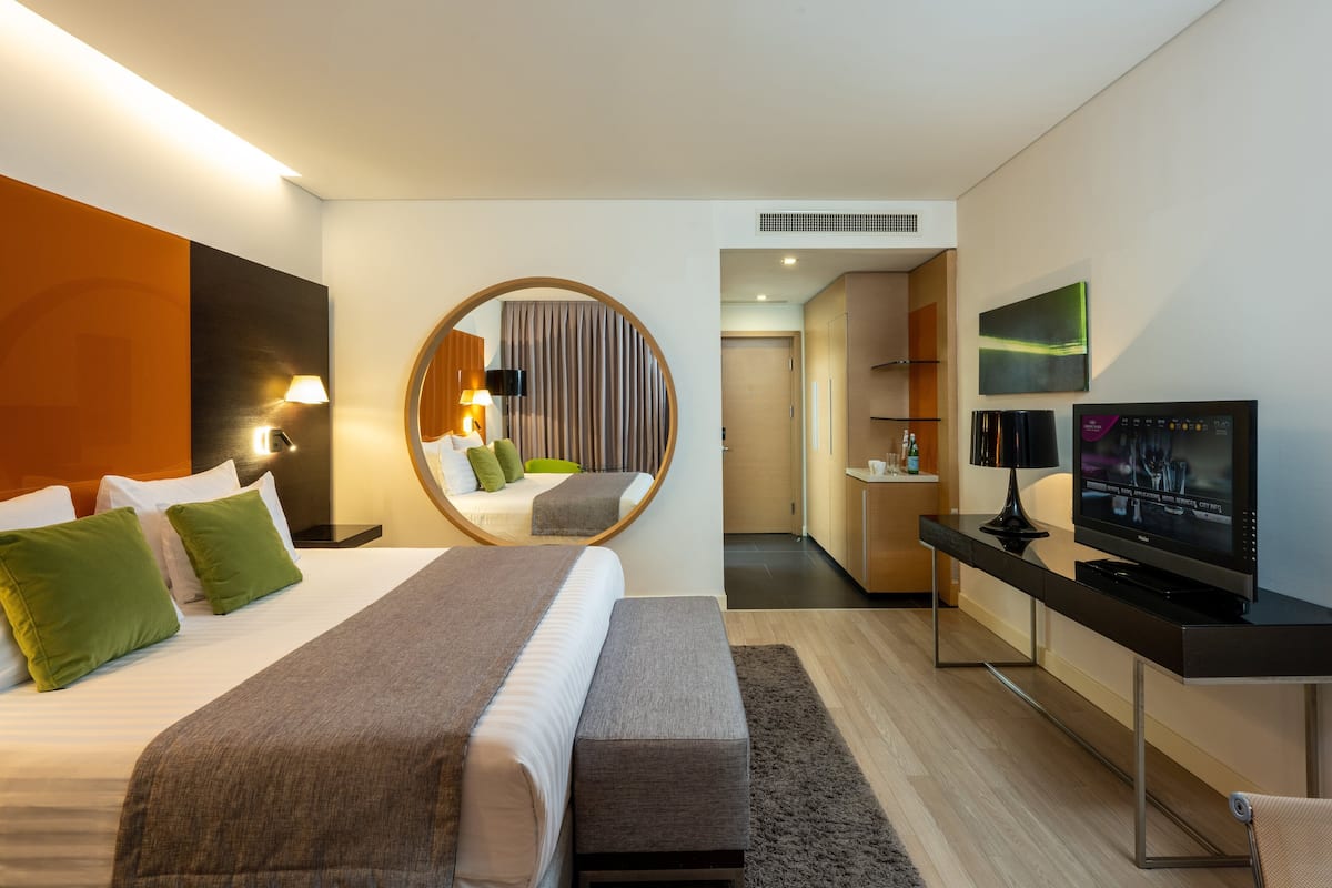Een moderne hotelkamer met een groot bed, een bureau met een flatscreen televisie, een ronde spiegel en een apart zitgedeelte.