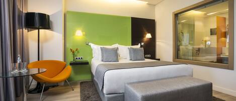 A modern hotel room with a large bed, a small table with glasses, an orange chair, and a large mirror.