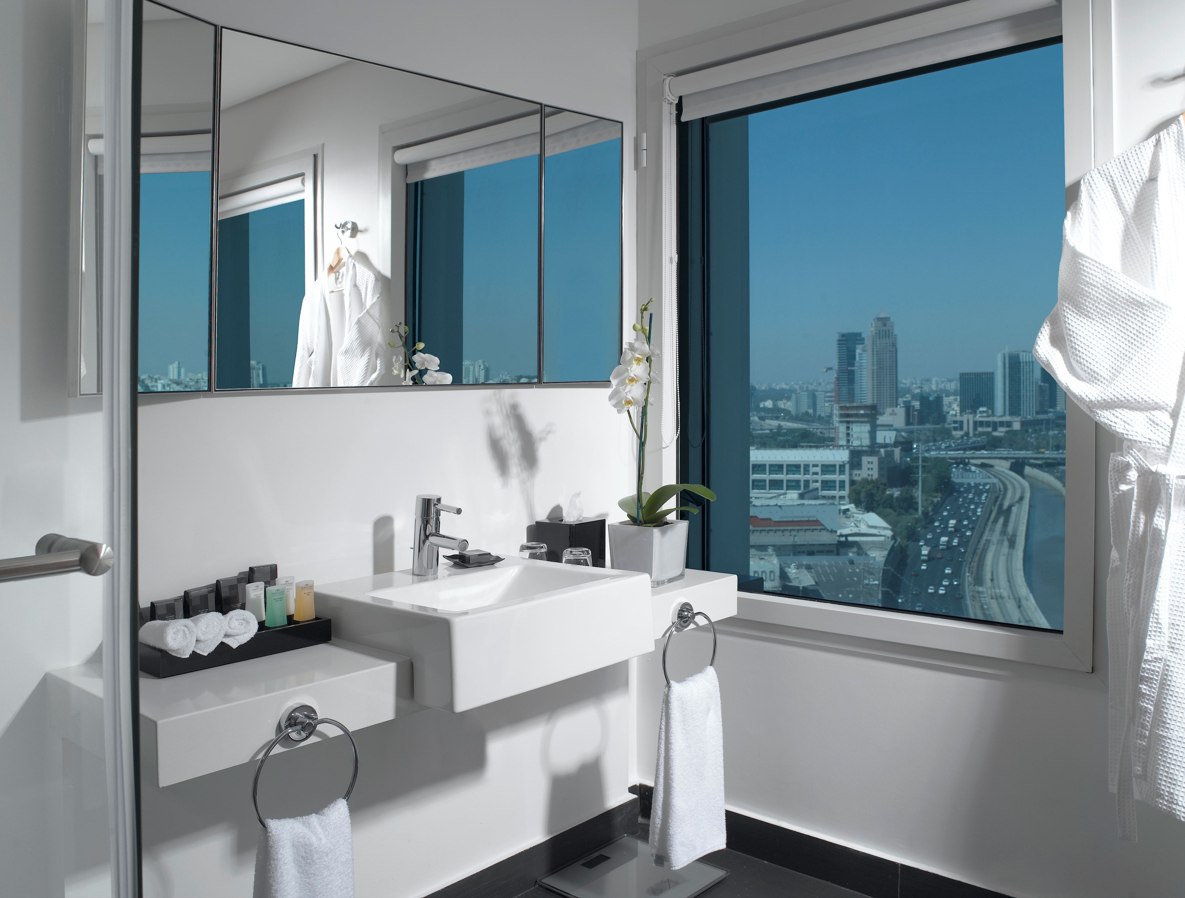 Une salle de bain moderne avec un grand miroir, un lavabo blanc et une vue sur le paysage urbain, visible à travers une grande fenêtre.