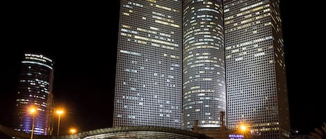 A nighttime view of a cityscape with modern high-rise buildings, including a prominent building with a grid-like facade and illuminated signage.