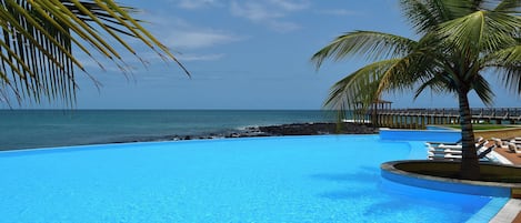 A clear blue pool with a wooden deck, palm trees, and a view of the ocean.