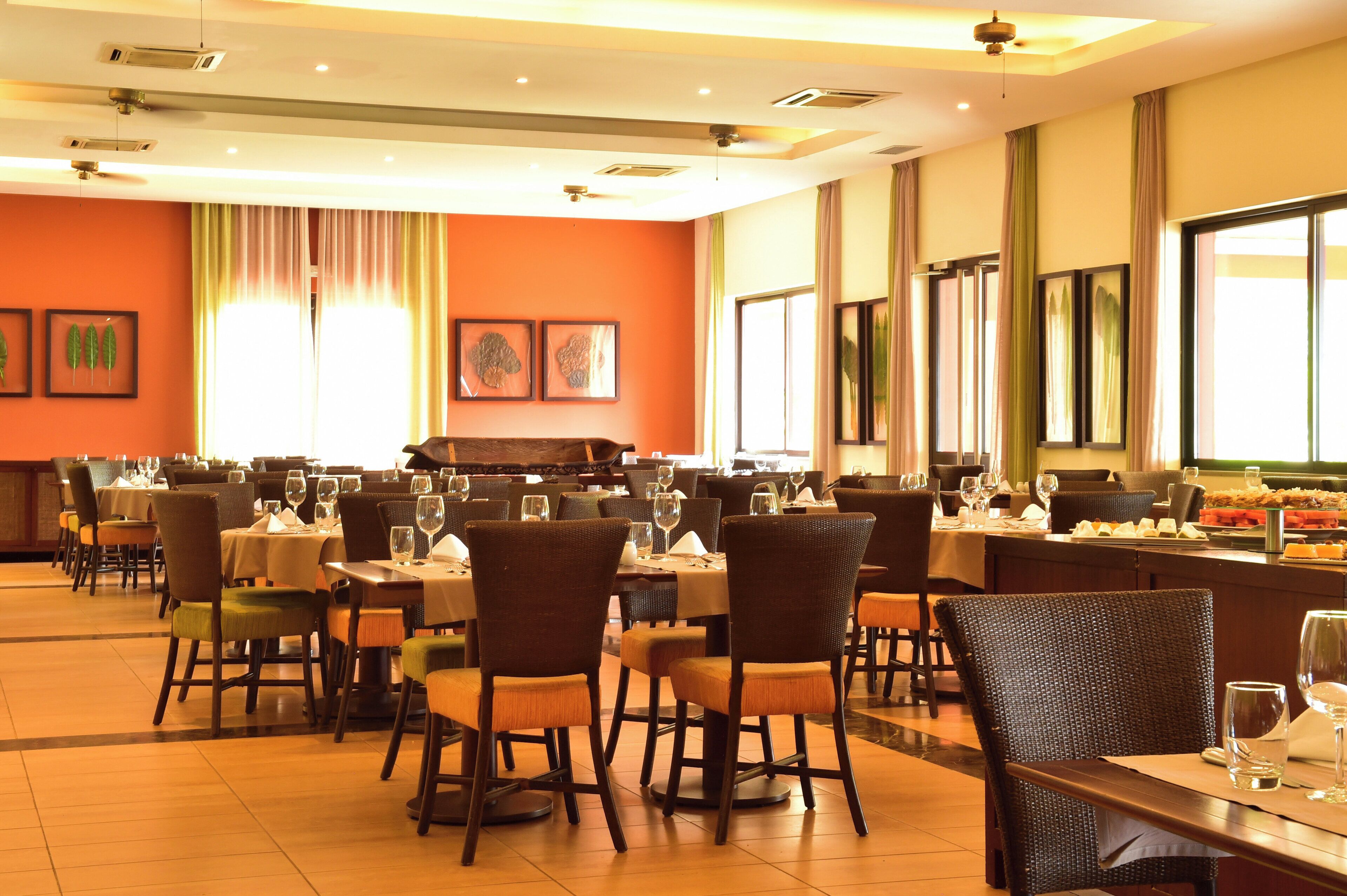 A restaurant with multiple dining tables and chairs, large windows with curtains, and framed artwork on the walls.