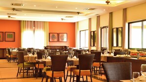 Een restaurant met meerdere eettafels en stoelen, grote ramen voorzien van gordijnen en kunstwerken in lijsten aan de muren.