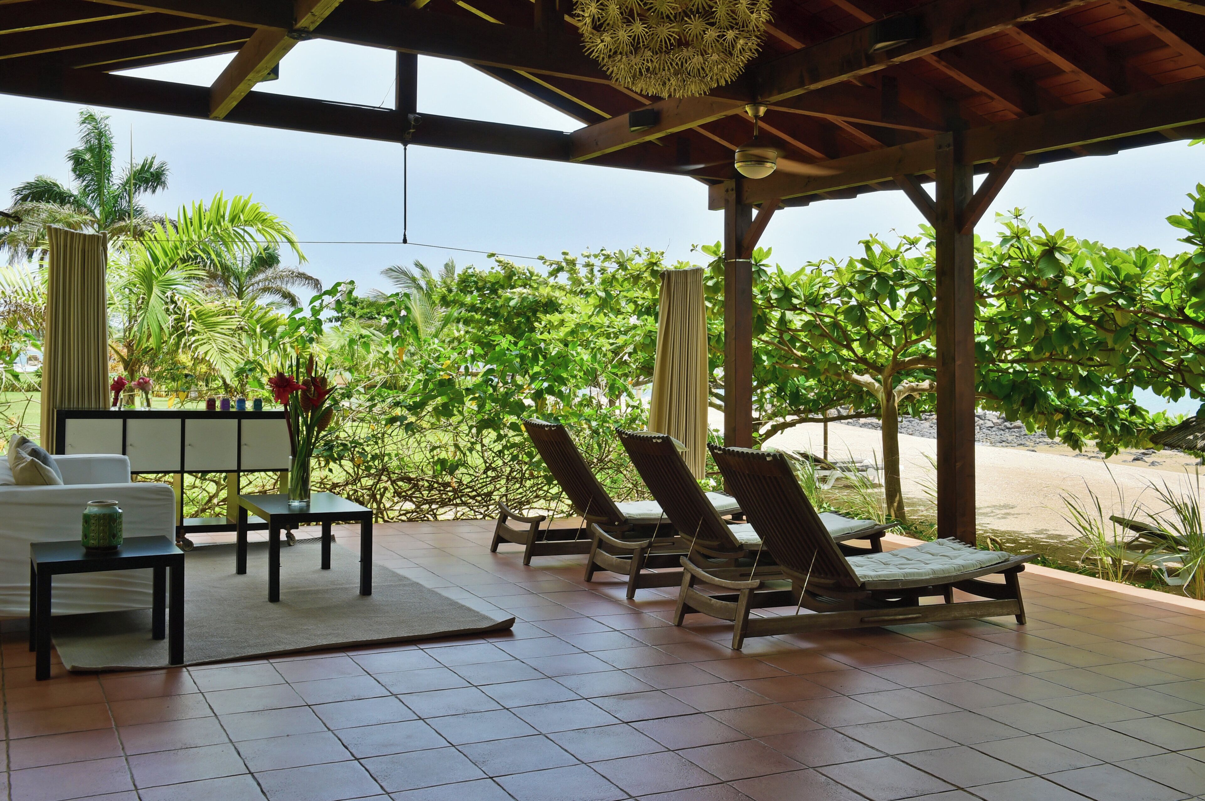 Un espace extérieur aménagé, abrité et doté de chaises longues et d’une table, offrant une vue sur une plage et une végétation luxuriante.