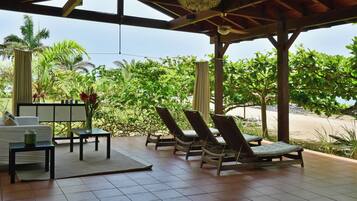 Un espace extérieur aménagé, abrité et doté de chaises longues et d’une table, offrant une vue sur une plage et une végétation luxuriante.