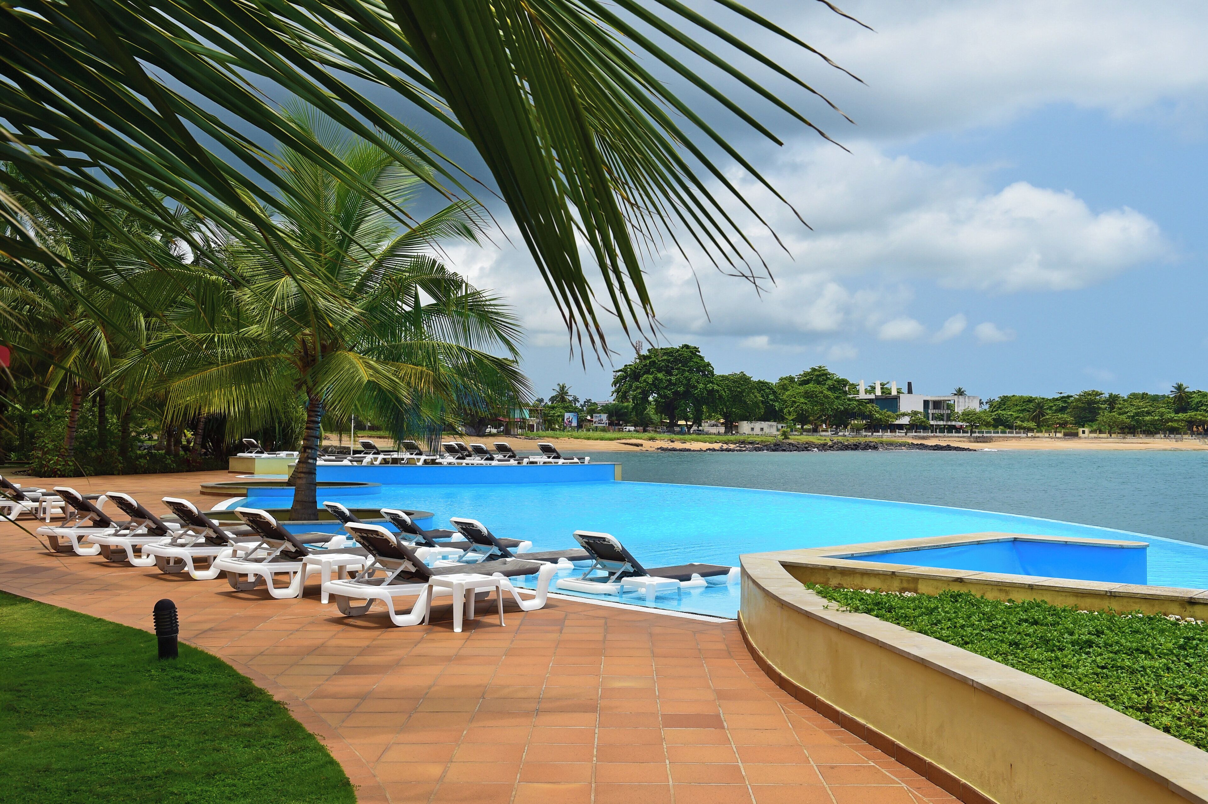 A pool area with lounge chairs and palm trees, overlooking a body of water and a beach.