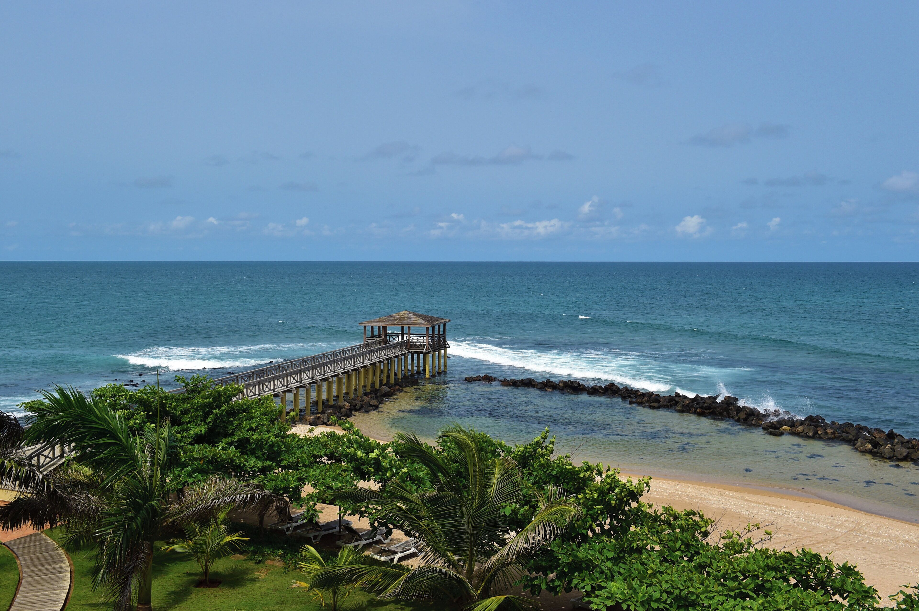 A beach with a wooden pier leading to a gazebo, surrounded by lush greenery and clear blue waters.