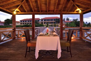 Una terraza de madera con una mesa lista para comer, con vista a un edificio de techo rojo y palmeras.