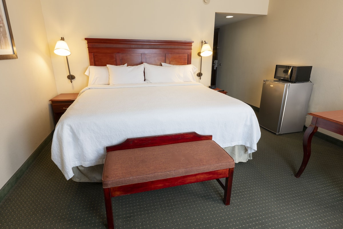 Une chambre d’hôtel avec un grand lit, deux lampes de chevet, une petite table, un banc et un réfrigérateur.