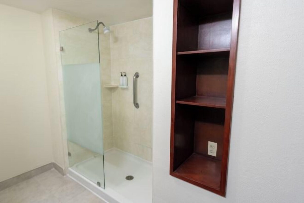 A bathroom with a glass shower enclosure, a wooden shelf, and a white wall.