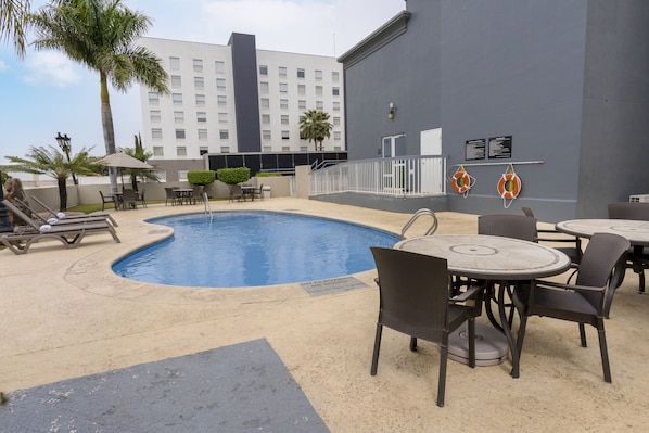 Un área de piscina con sillas de descanso, mesas y un edificio al fondo.