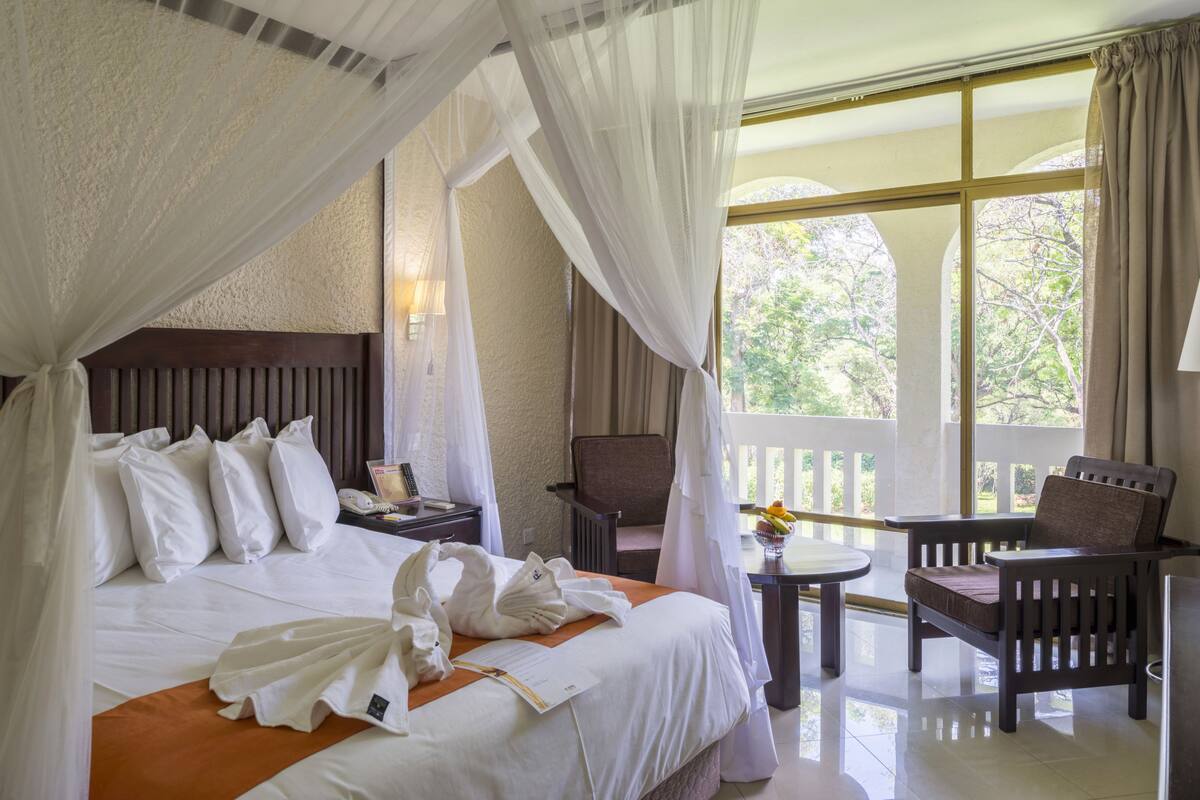 A hotel room with a bed, a chair, a small table, and a balcony with a view of trees.