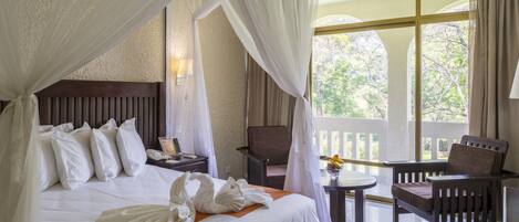A hotel room with a bed, a chair, a small table, and a balcony with a view of trees.