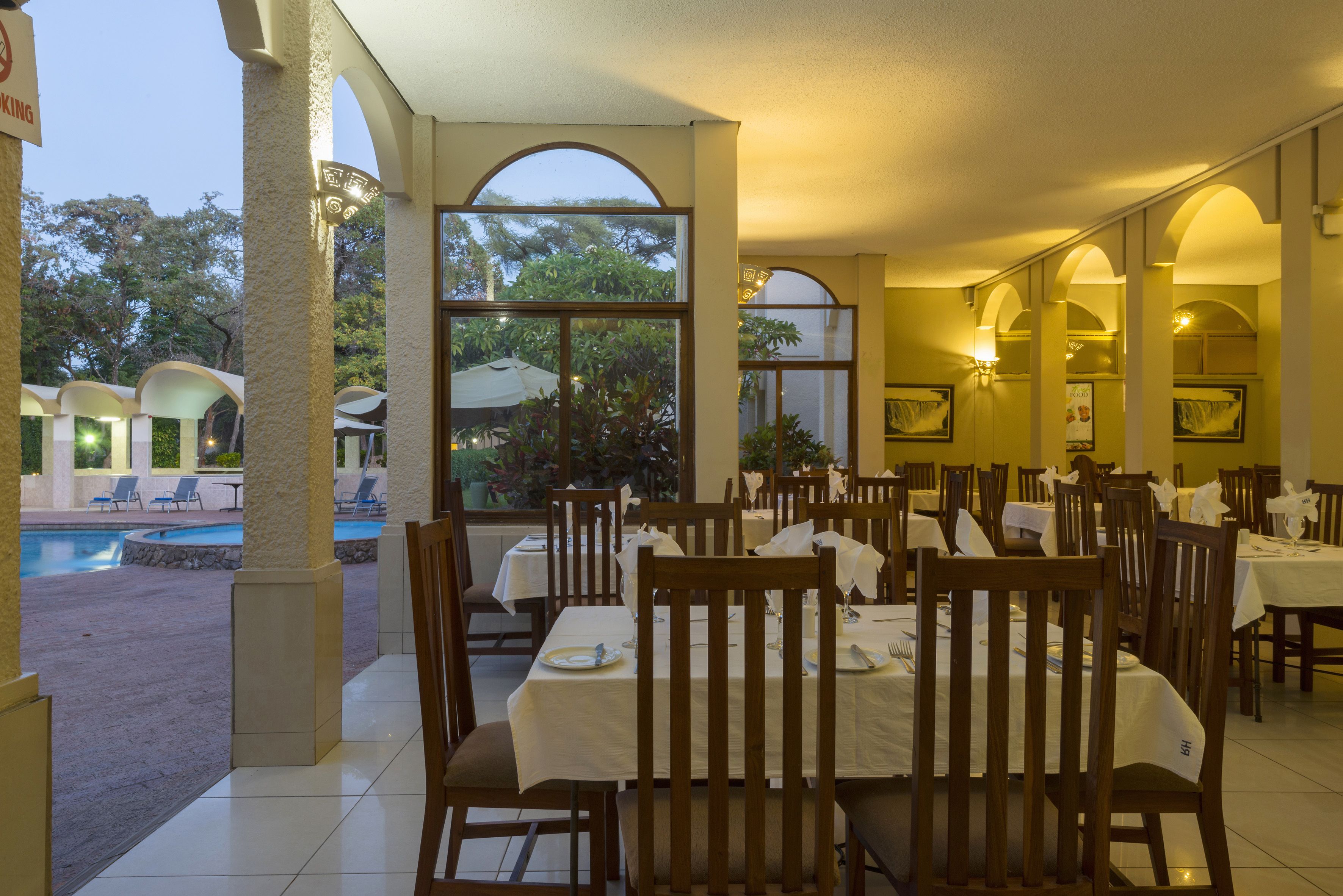 A restaurant with outdoor seating overlooking a pool and greenery. Tables are set with white tablecloths and wooden chairs.