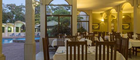 A restaurant with outdoor seating overlooking a pool and greenery. Tables are set with white tablecloths and wooden chairs.