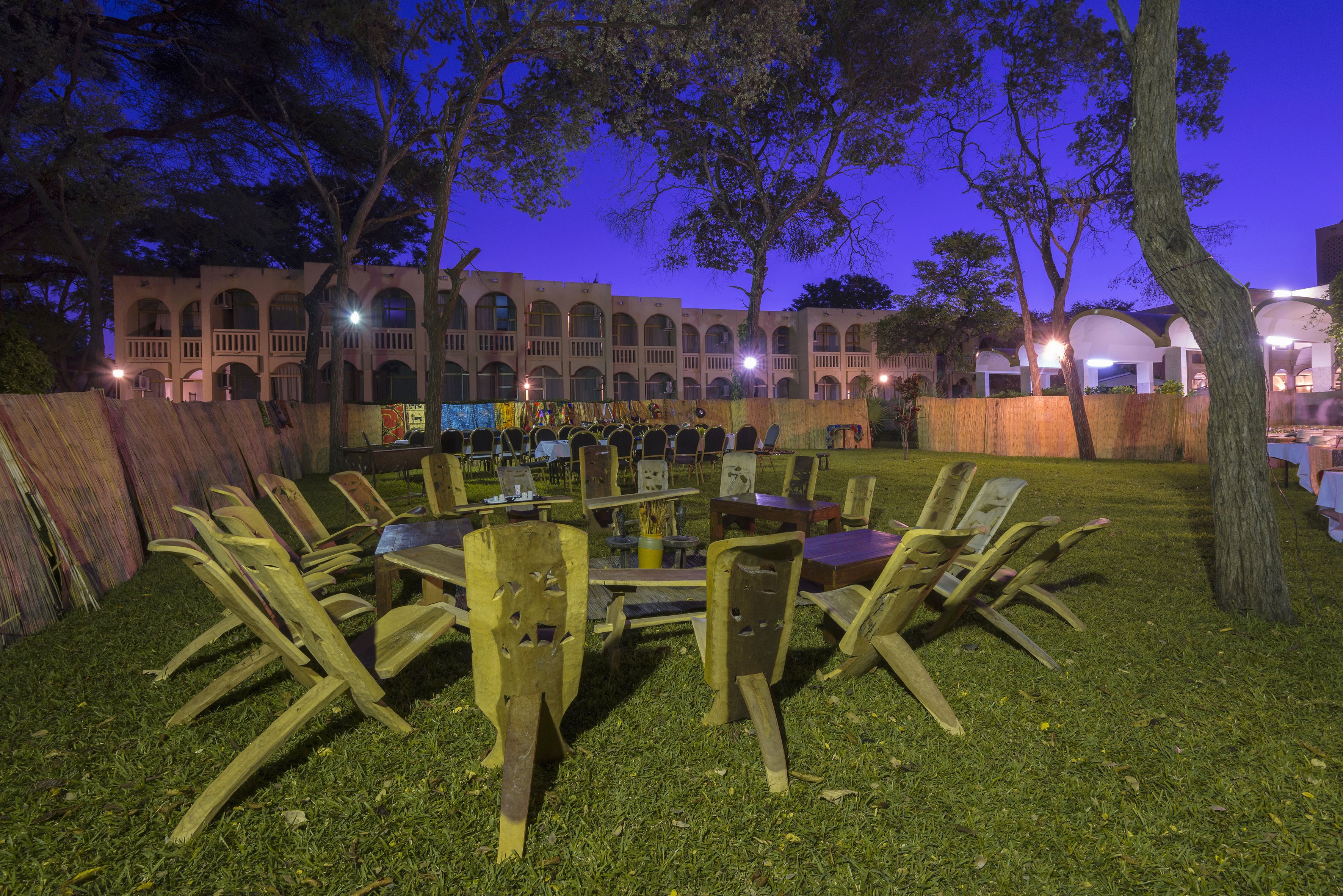 An outdoor dining area with a round table and chairs, surrounded by a grassy lawn and trees, with a building in the background.