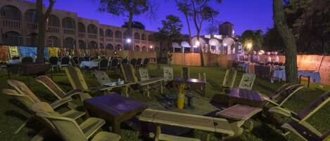 An outdoor seating area with wooden furniture and tables, surrounded by trees and a building with arched windows in the background.