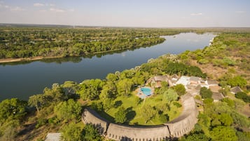 Explore a historic fort surrounded by lush greenery and a serene lake, with traditional thatched-roof buildings and a swimming pool.