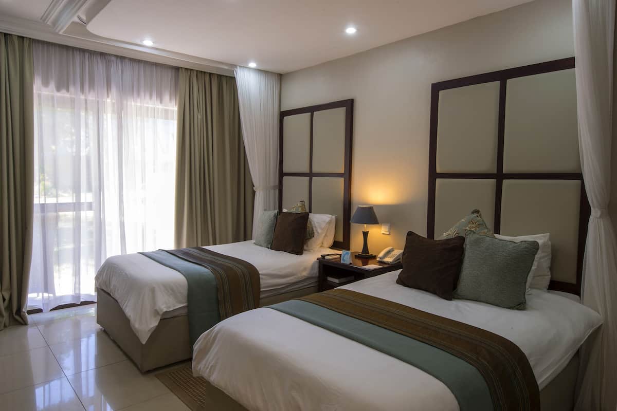 Ein Hotelzimmer mit zwei Betten, einem Nachttisch mit Lampe und einem großen Fenster mit Vorhängen.