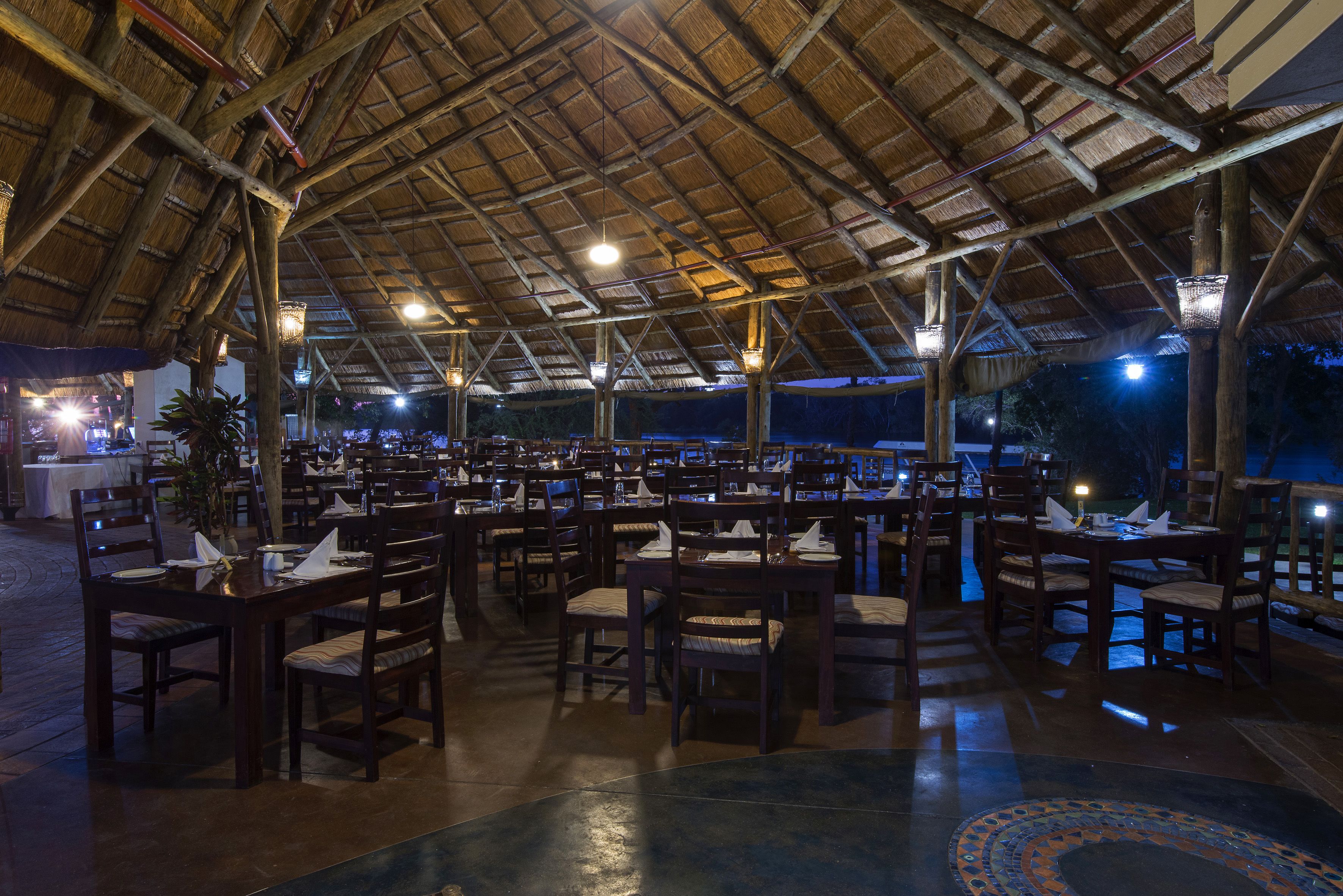 An indoor dining area with multiple tables and chairs, a thatched roof, and hanging light fixtures.