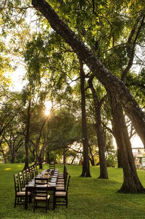 An outdoor dining area with a long table set for six, surrounded by chairs, under large trees with sunlight filtering through. - A'Zambezi River Lodge (Victoria Falls)