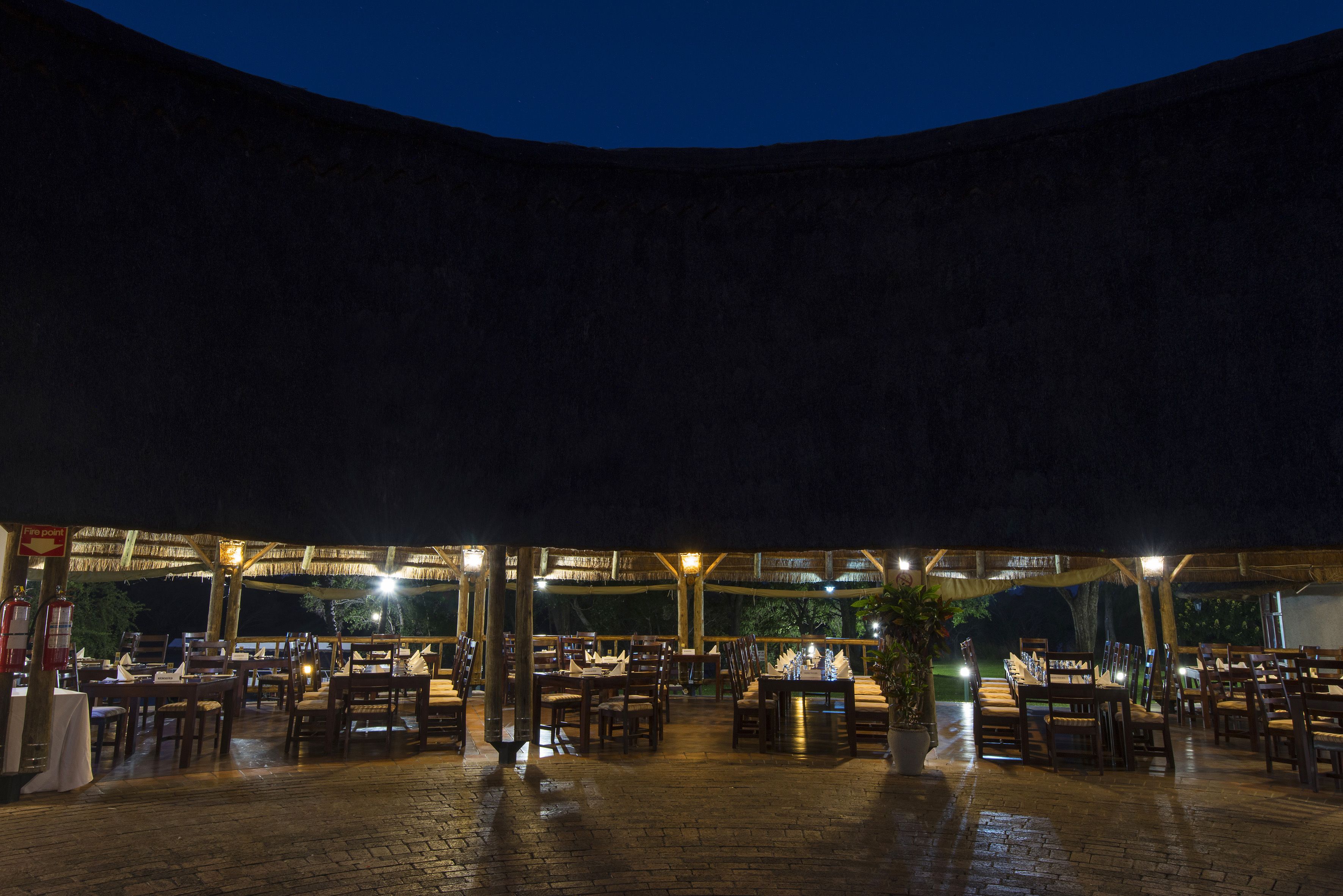An outdoor restaurant with multiple tables and chairs, covered by a wooden structure with lights.