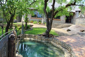 A stone pool surrounded by a brick pathway, with a stone building in the background. - Lodge at The Ancient City (Masvingo)