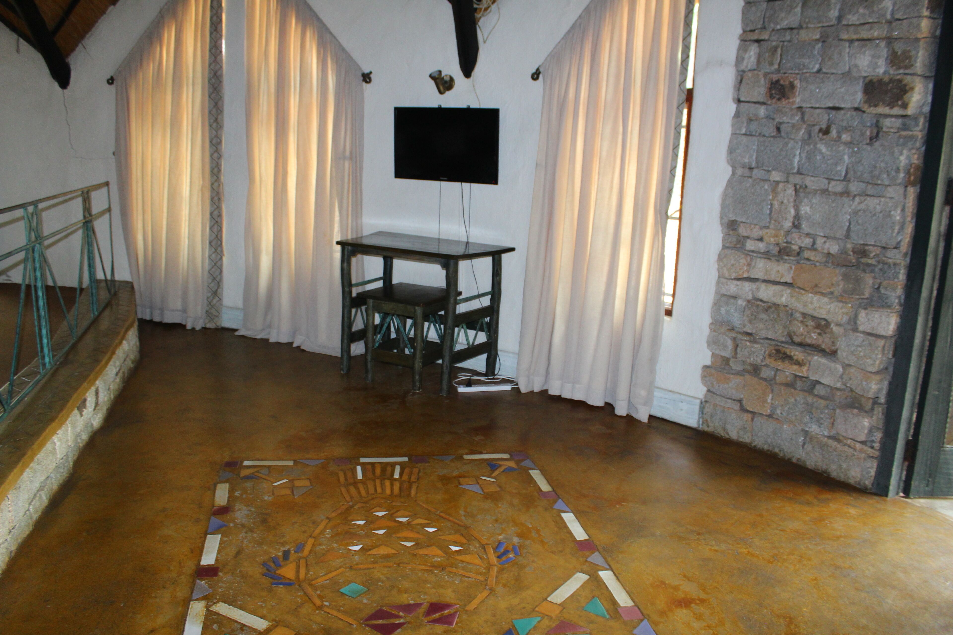 A room with a tiled floor, a table with chairs, a mounted TV, and a stone wall.