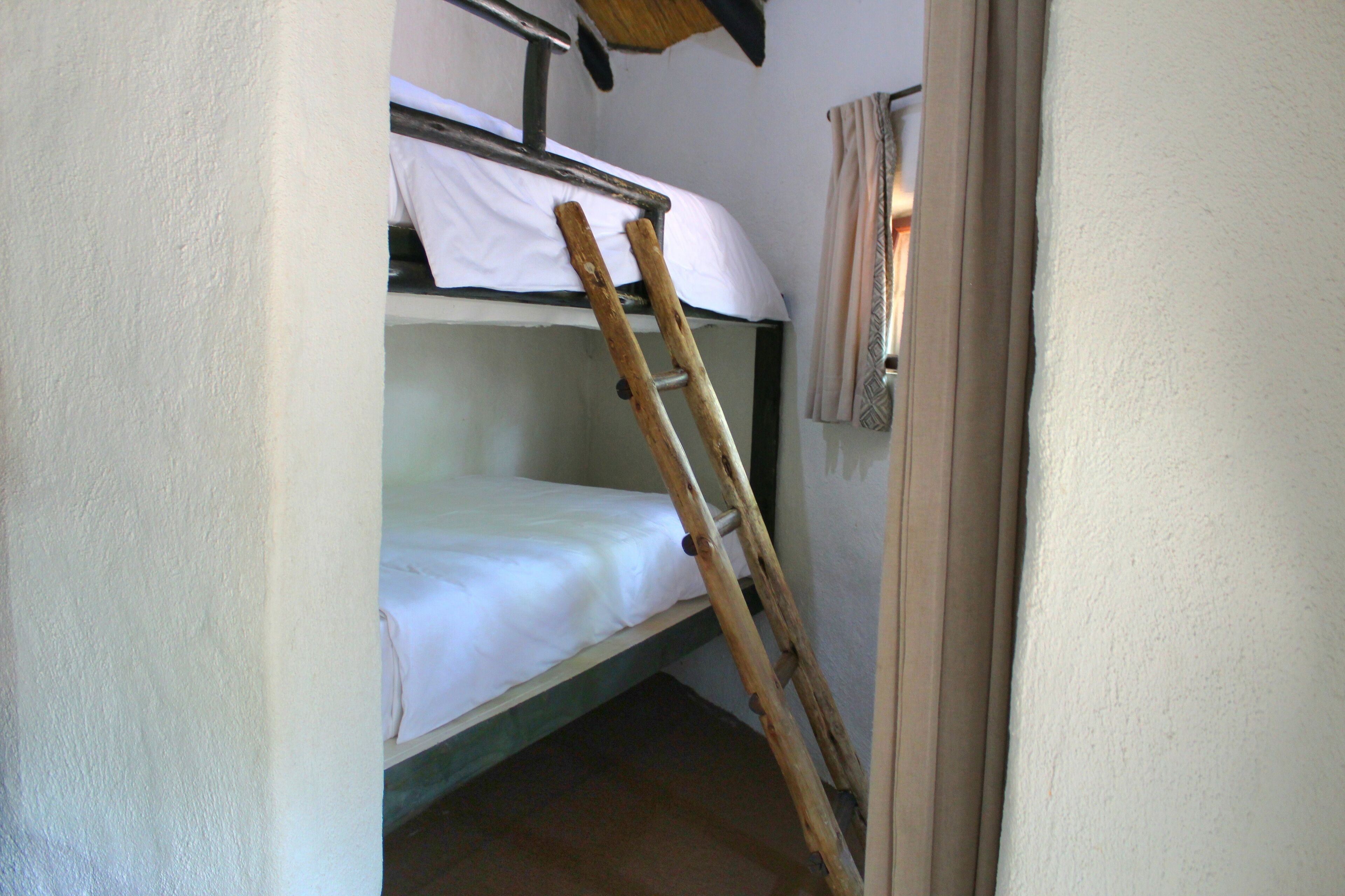 Ein Stockbett mit einer Holzleiter, die dagegenlehnt, ein weißes Laken auf der unteren Etage und ein Vorhang am Fenster.