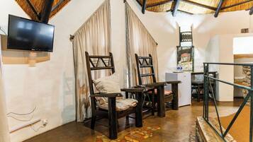 A room with a TV mounted on the wall, two wooden chairs, a small table, and a thatched roof ceiling.