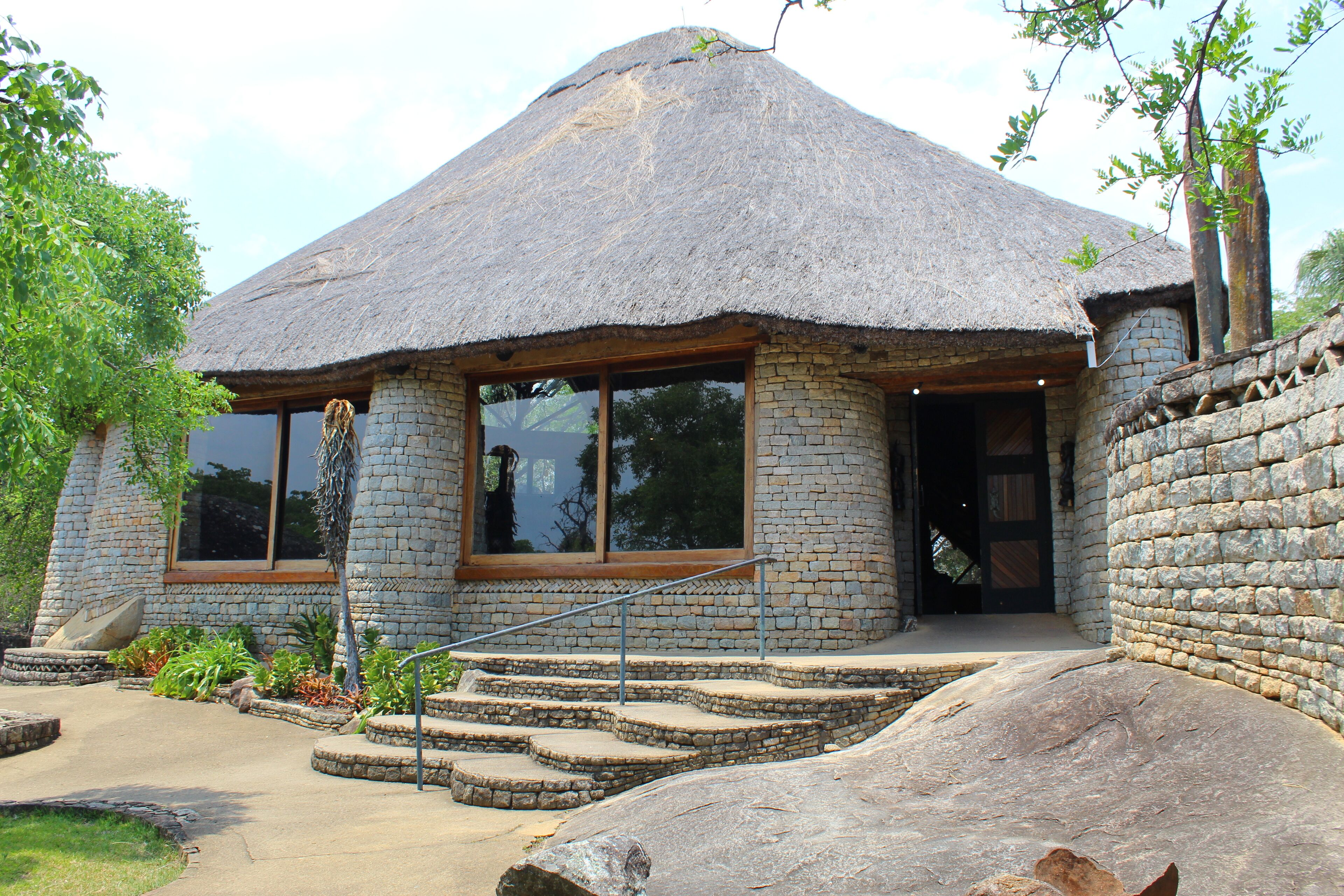 A stone building with a thatched roof, large windows, and a stone staircase leading to the entrance.