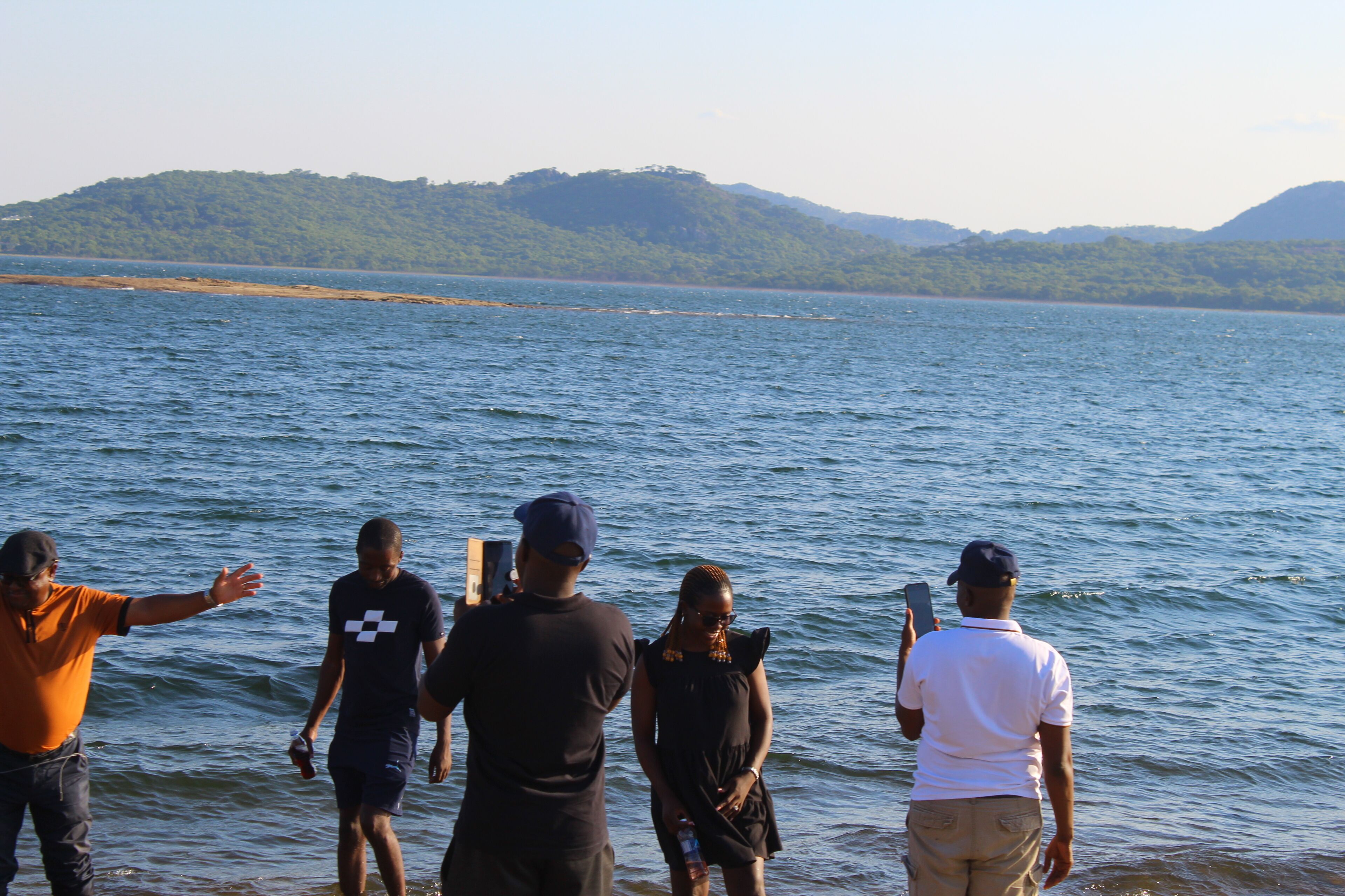 Un groupe de personnes se tient près d’un lac, avec un paysage vallonné en arrière-plan.