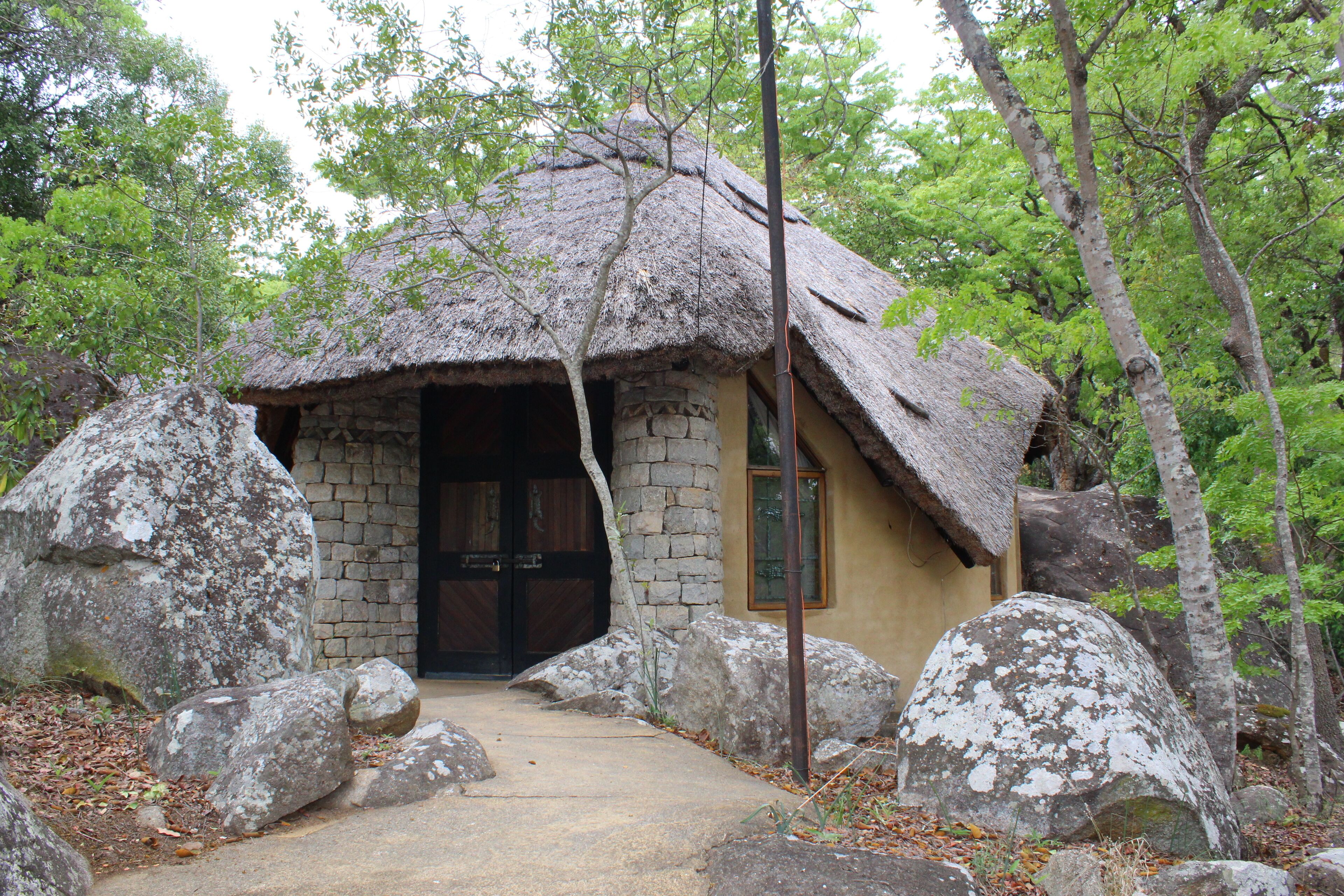 A rustic stone building with a thatched roof, surrounded by large rocks and trees.