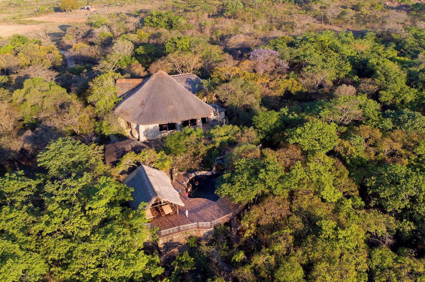 Uma pousada com um edifício principal coberto de palha e uma construção menor com telhado semelhante, cercada por floresta densa.