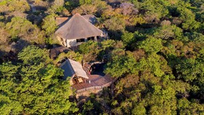 A lodge with a thatched-roof main building and a smaller structure with a similar roof, surrounded by dense forest. - Lodge at The Ancient City (Masvingo)