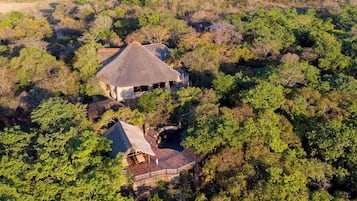 Uma pousada com um edifício principal coberto de palha e uma construção menor com telhado semelhante, cercada por floresta densa.