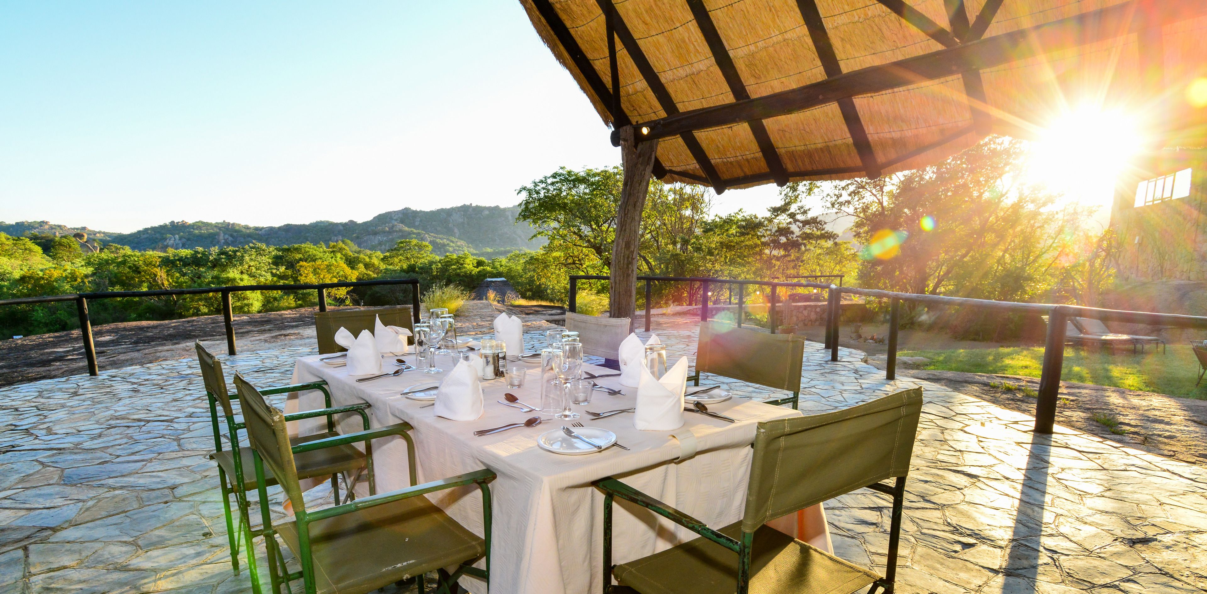 An outdoor dining area with a table set for two, surrounded by stone flooring and green chairs, under a wooden pergola with a view of a mountainous landscape.