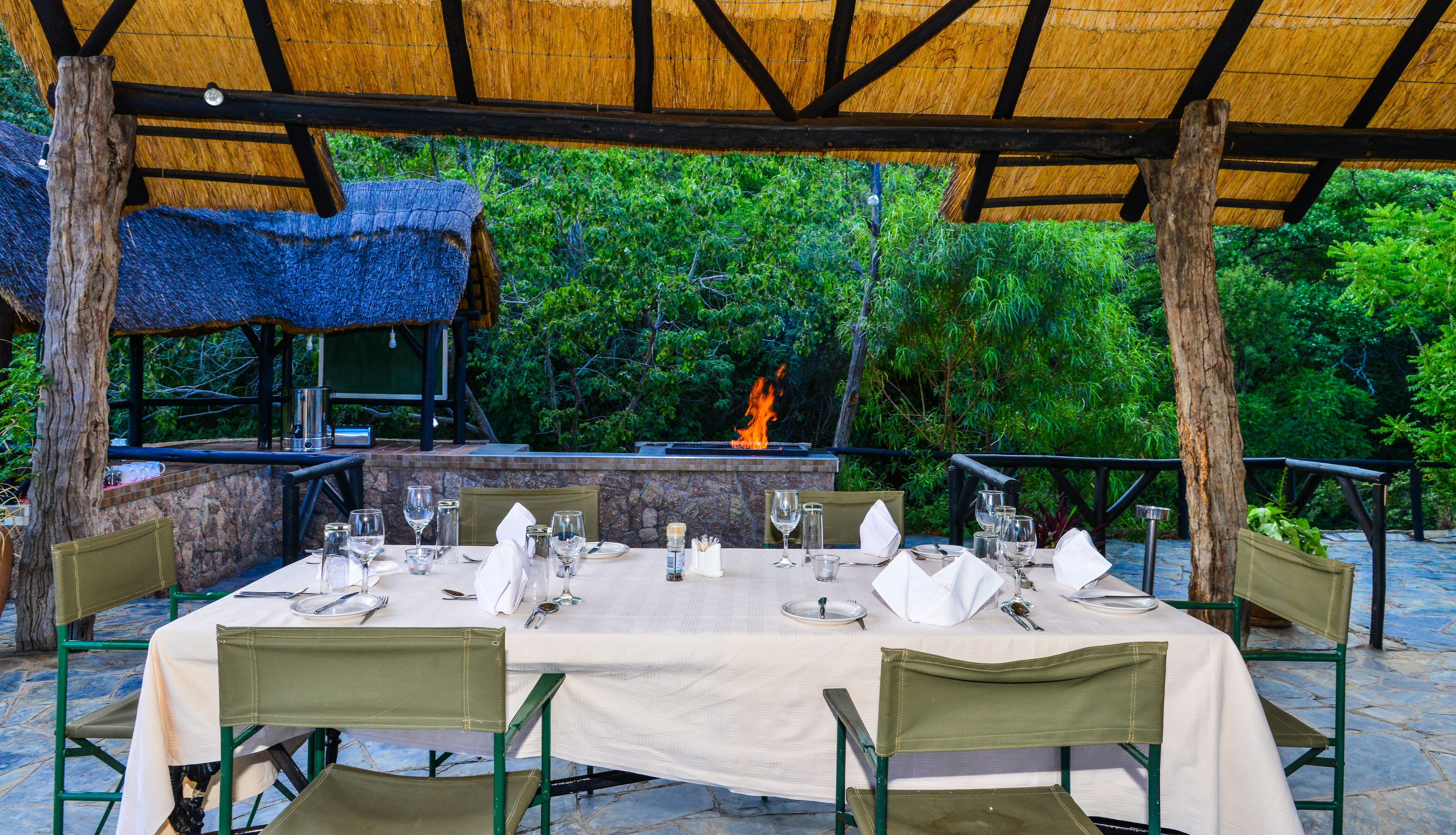An outdoor dining area with a stone fireplace, green chairs, and a thatched roof structure.