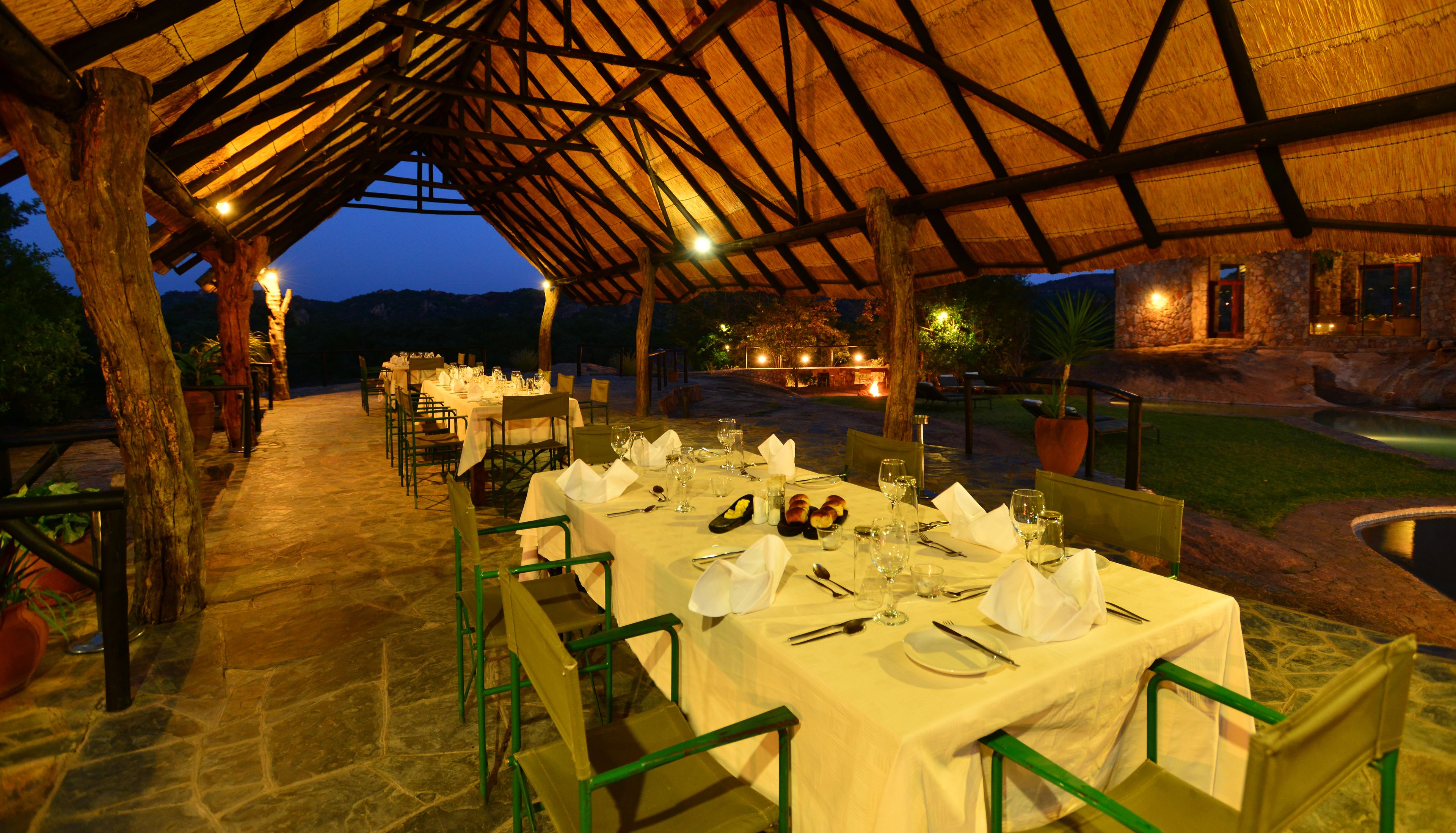 An outdoor dining area with tables set for a meal, under a wooden pergola with a thatched roof.