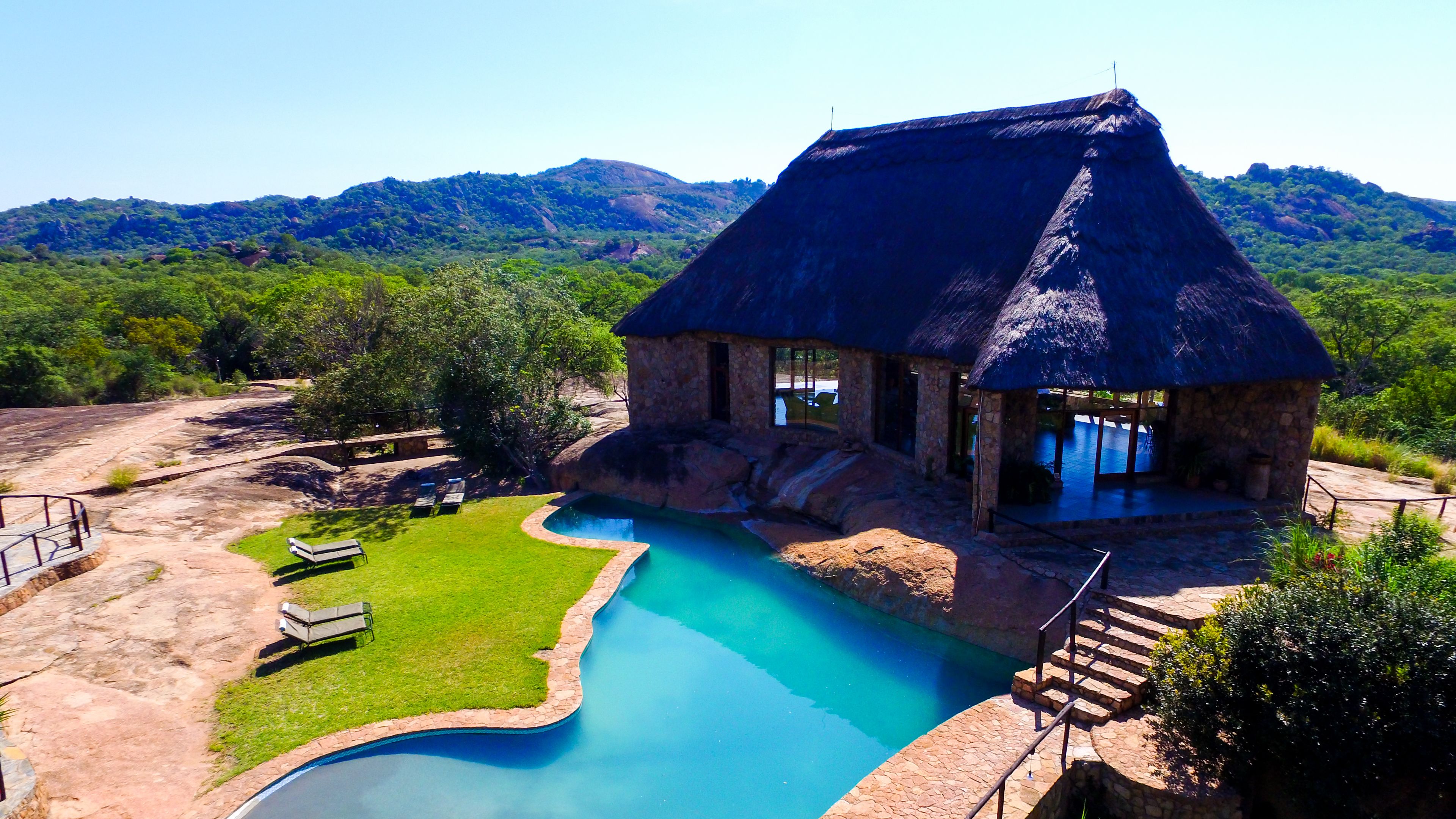 A thatched-roof building with a swimming pool and a grassy area with lounge chairs.