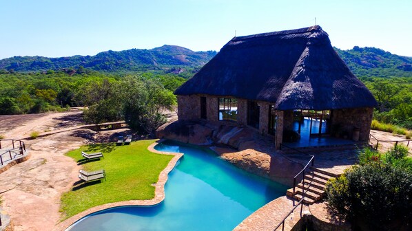 A thatched-roof building with a swimming pool and a grassy area with lounge chairs.