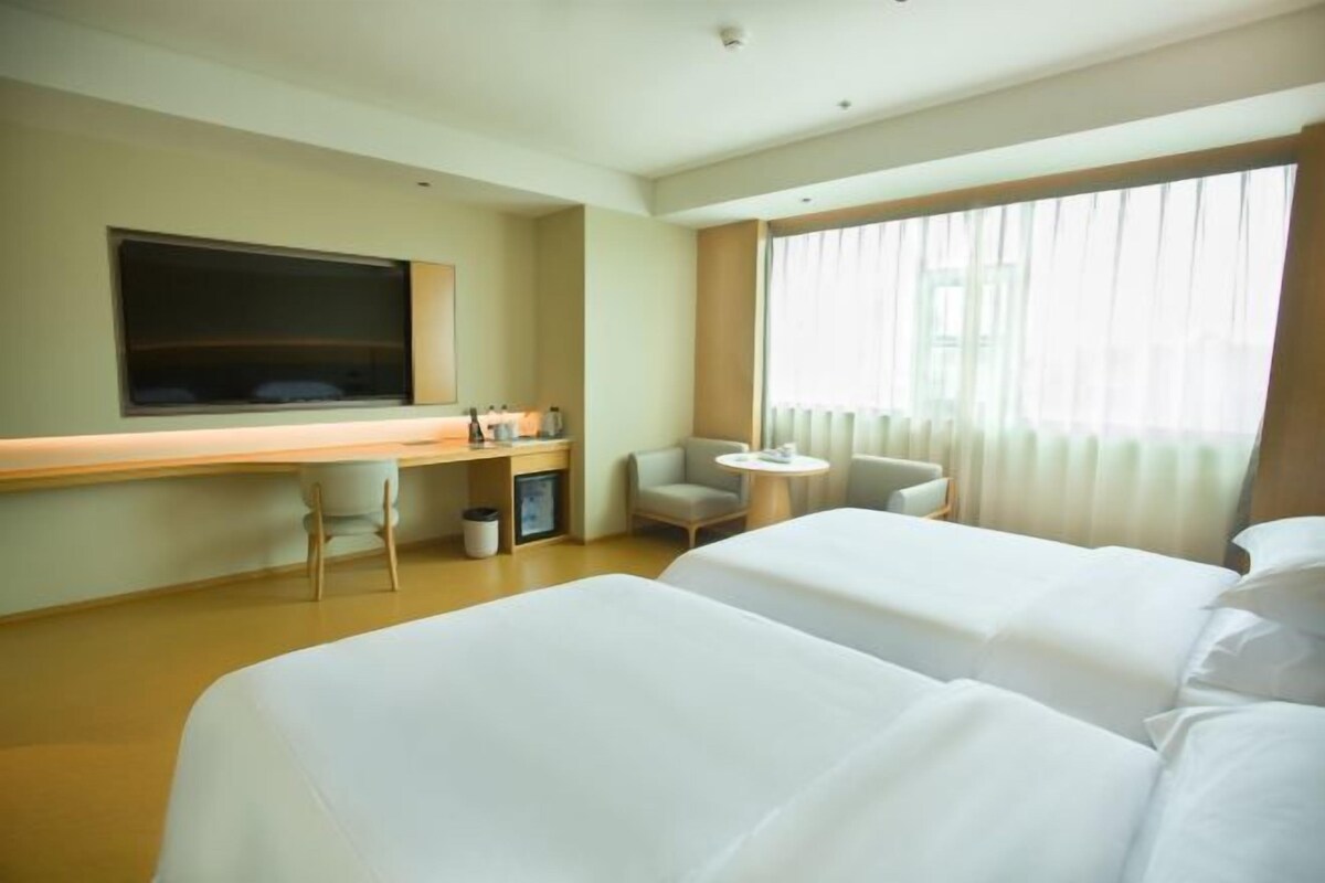 Habitació d'hotel amb dos llits, una taula de treball, una cadira, un televisor i una finestra amb cortines.