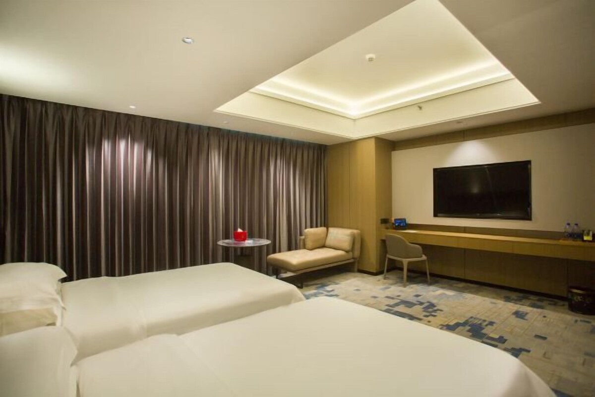 Quarto de hotel com duas camas, uma escrivaninha, uma cadeira, televisão e uma janela grande.