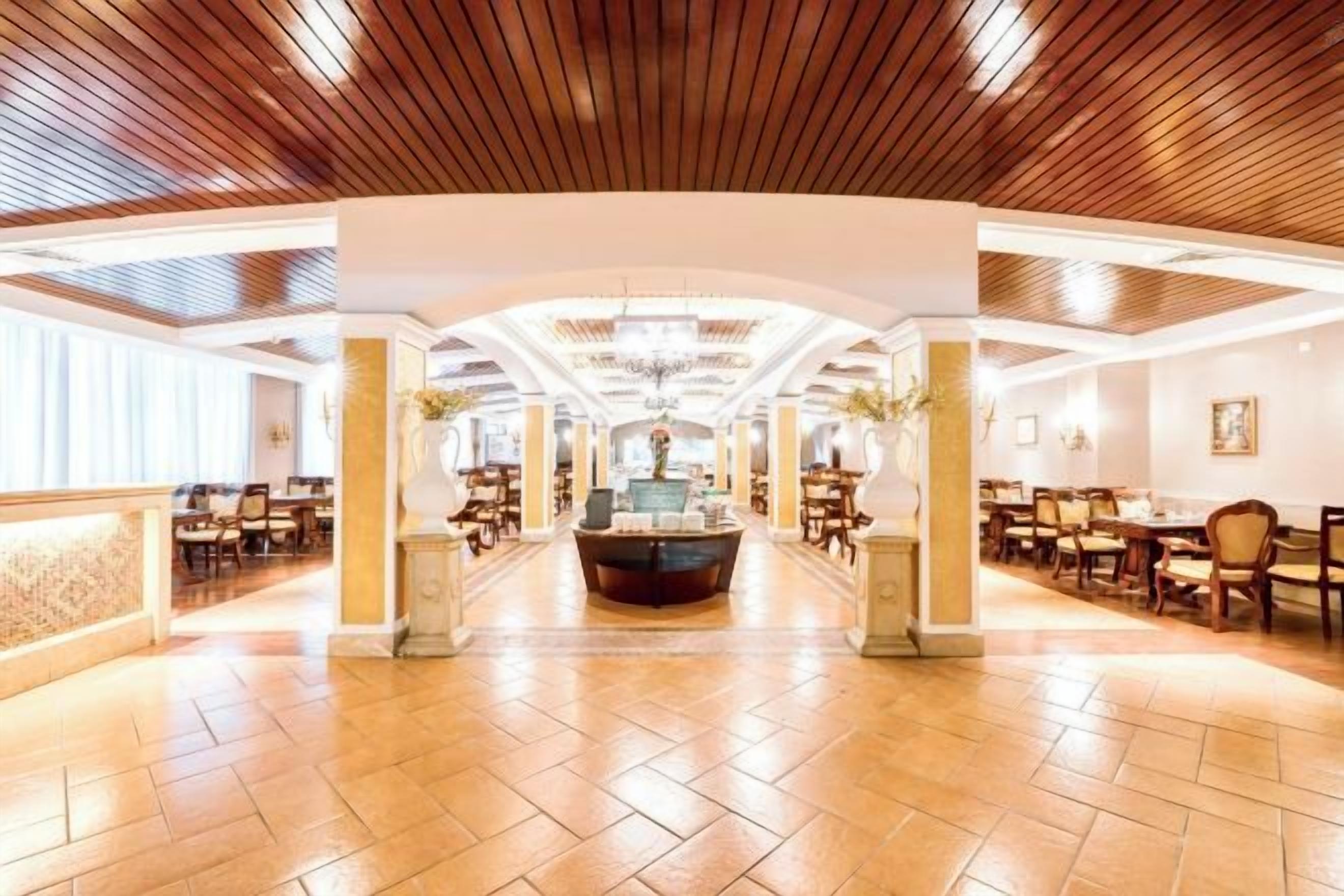 A spacious hotel lobby with a central reception desk, high ceilings with wooden paneling, and a tiled floor.