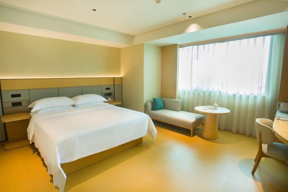 Quarto de hotel com uma cama grande, mesa de cabeceira, mesa redonda pequena e um espaço de estar com sofá e poltrona.