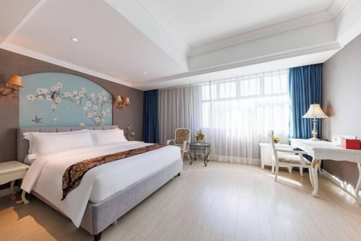 A hotel room with a large bed, a desk, a chair, a small table, and a decorative wall piece.