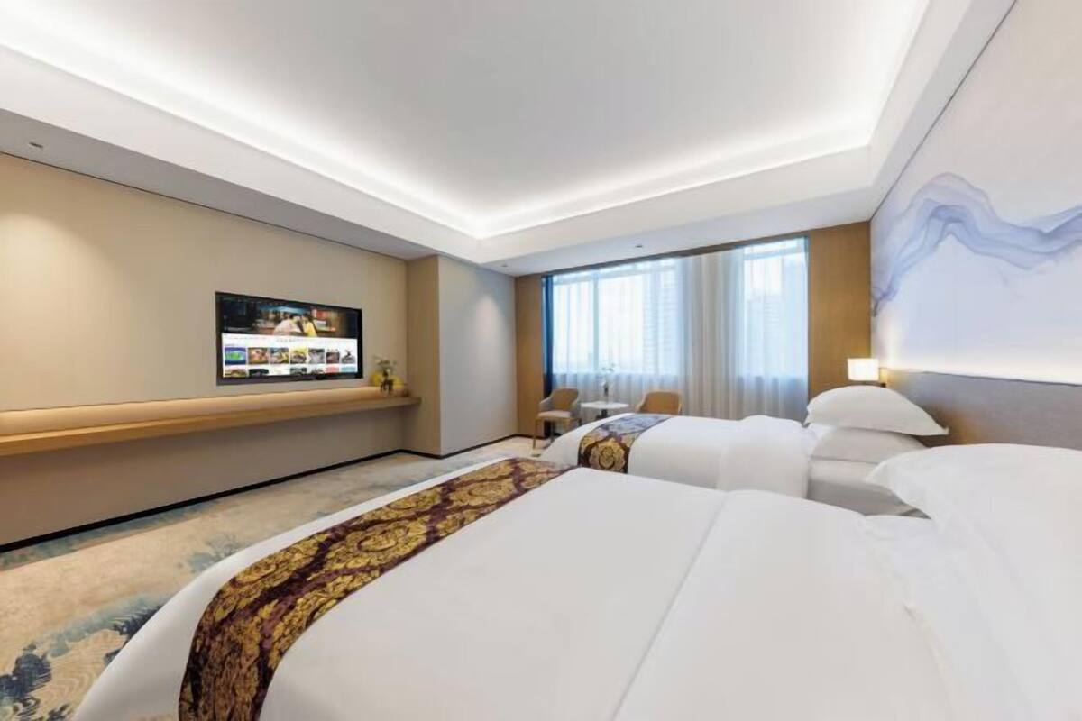 A hotel room with two beds, a TV on the wall, a desk, and a window with curtains.