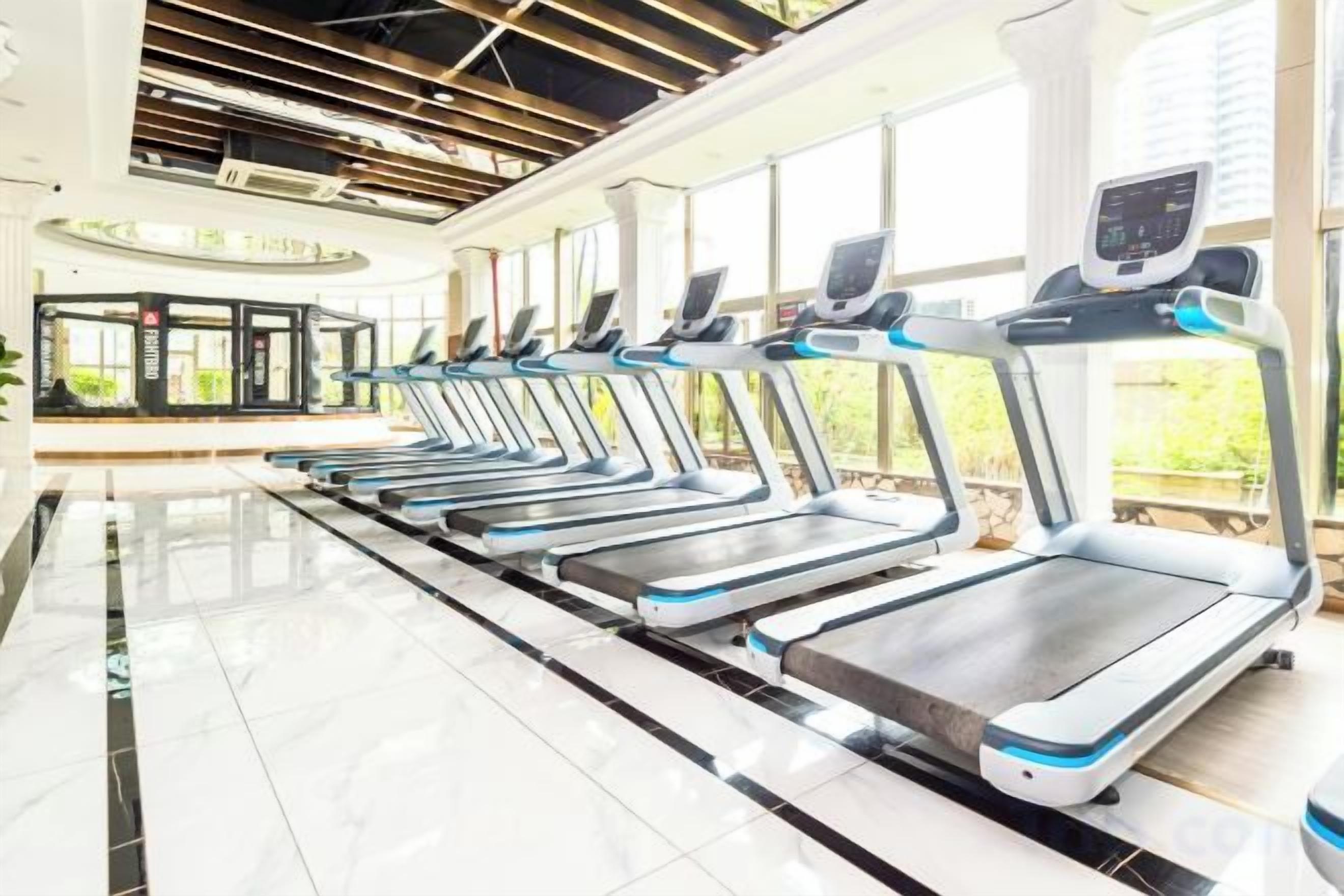 Une rangée de tapis de course dans une salle de sport au design moderne, dotée de grandes fenêtres et d’un sol carrelé et impeccable.